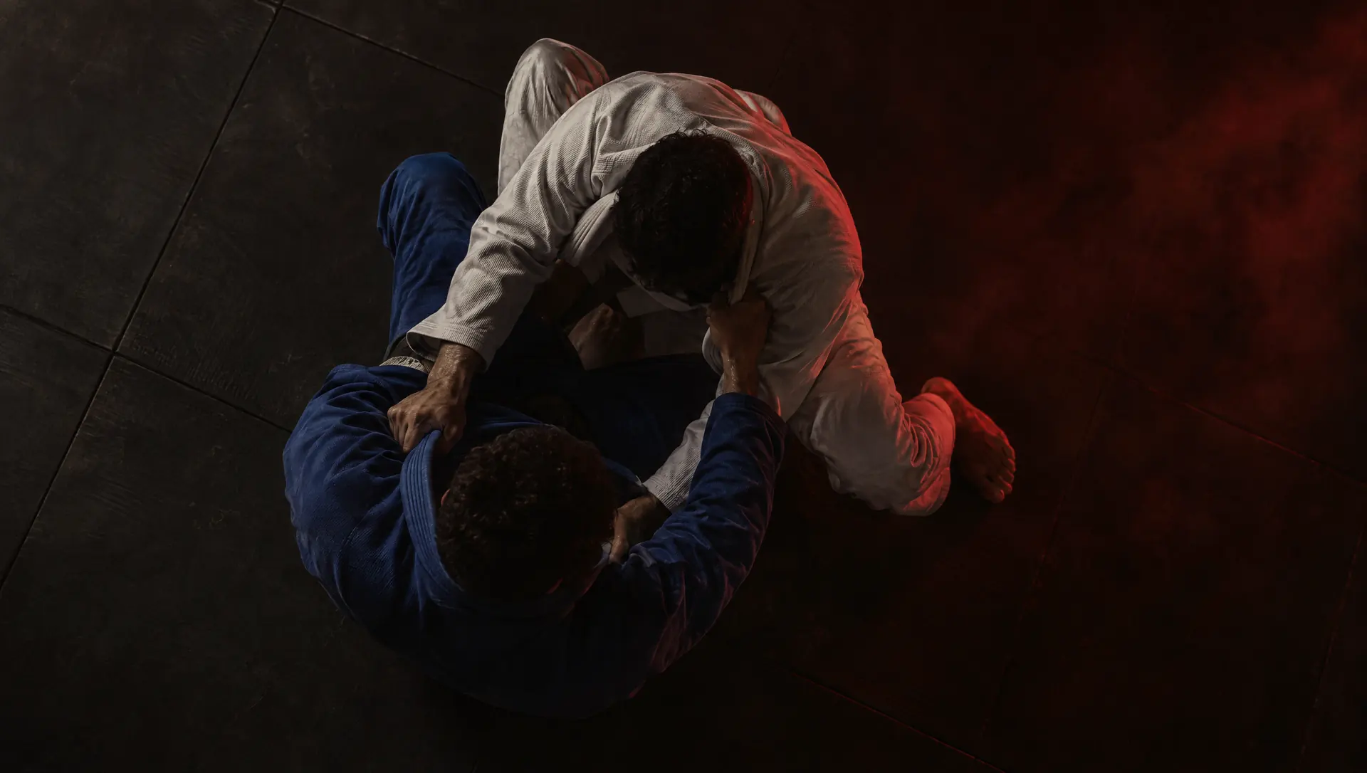 Two grapplers in white and blue gis, seen from above, working for collar grips on dark mats.