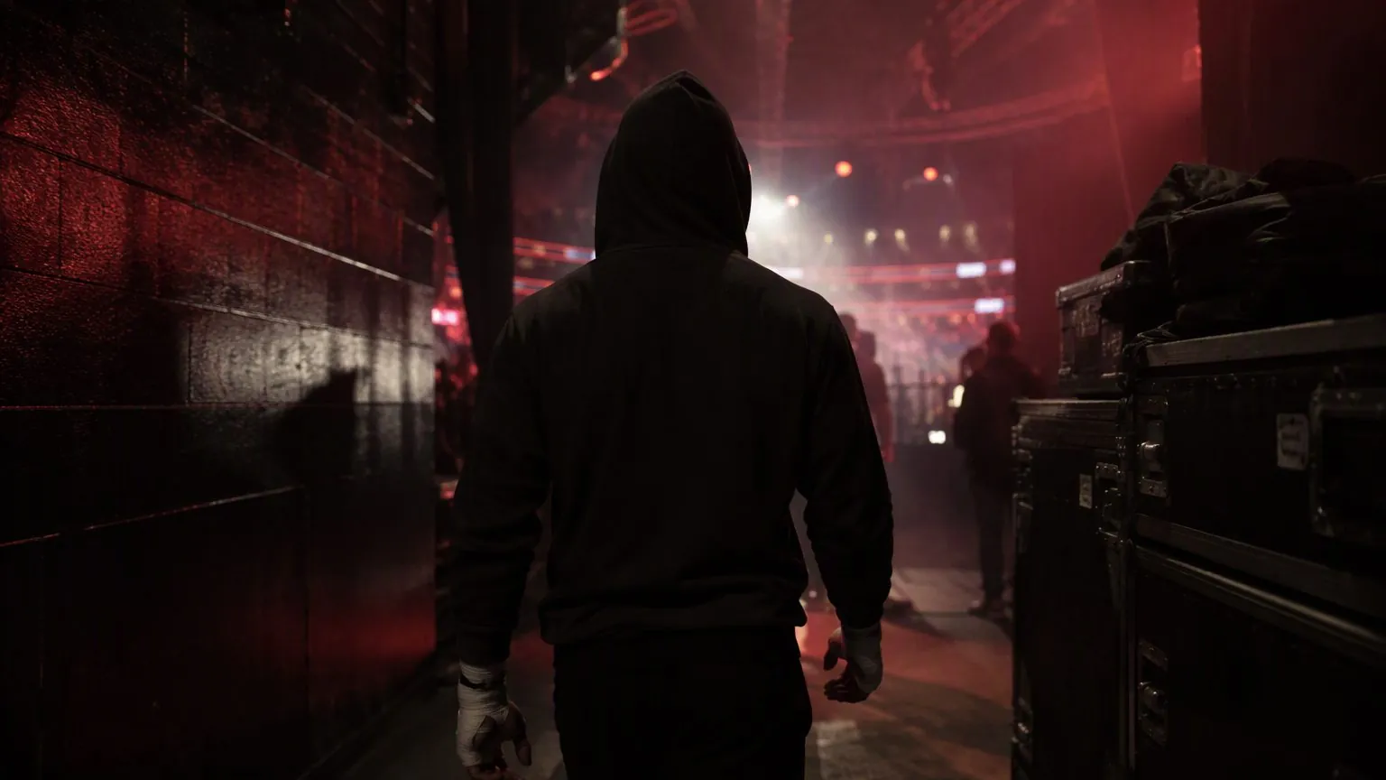 A hooded fighter with wrapped hands walks down the tunnel toward the arena lights.